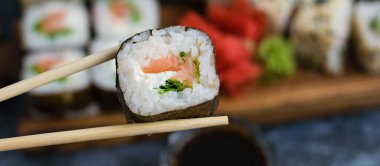 Elleri ahşap çubuklu suşi rulosu ıspanak, peynir, somon ve nori. Arka planda büyük rulo seti.