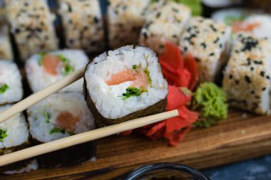 Elleri ahşap çubuklu suşi rulosu ıspanak, peynir, somon ve nori. Arka planda büyük rulo seti.