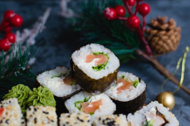 Ahşap bir tahtada büyük bir suşi seti. Susam tohumlu rulo, nori, peynir, kırmızı balık, salatalık ve yılan balığı, zencefil, wasabi ve gri arka planda noel dekoru.