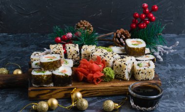 Susam tohumlu rulo, nori, peynir, kırmızı balık, salatalık ve yılan balığı, zencefil, wasabi, soya sosu ve gri arka planda Noel dekoru. Tahtada büyük bir suşi seti.