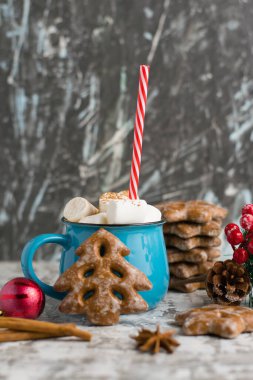 Kırmızı plastik tüp, lezzetli kurabiyeler, tarçın çubukları, yıldız anasonu ve gri arka planda noel dekoru olan mavi bardakta tarçınlı ve marşmelovlu kakao içeceği..