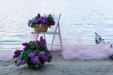 Nehir kenarında sepetin içinde leylak rengi bir buket. Pembe kumaştan, beyaz sandalyeden ve sepetteki çiçeklerden oluşan güzel bir dekor..