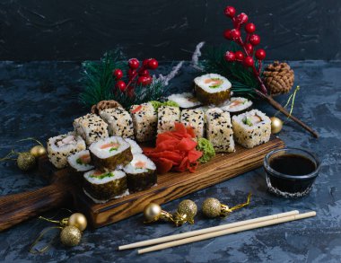 Ahşap bir tahtada büyük bir suşi seti. Susam tohumlu rulo, nori, peynir, kırmızı balık, salatalık ve yılan balığı, zencefil, wasabi, soya sosu ve koyu arkaplanda noel dekoru.