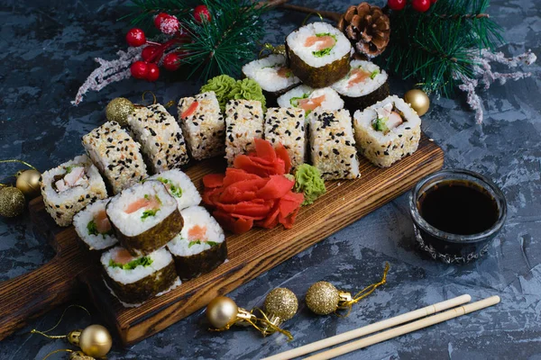 Large set of sushi on a wooden board top view. Rolls with sesame seeds, nori, cheese, red fish, cucumber and eel, ginger, wasabi, soy sauce, bamboo sticks and christmas decor on gray background.