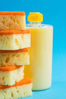 Şeffaf plastik bir şişede bulaşık deterjanı ve mavi arka planda gözenekli sarı temizlik süngeri. Ekolojik deterjan kavramı.