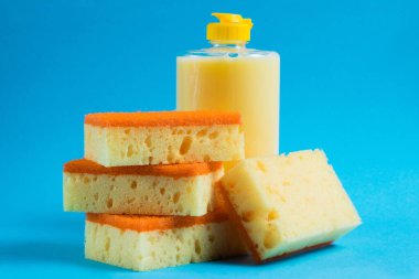 Şeffaf plastik bir şişede bulaşık deterjanı ve mavi arka planda gözenekli sarı temizlik süngeri. Ekolojik deterjan kavramı.