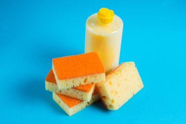 Şeffaf plastik bir şişede bulaşık deterjanı ve mavi arka planda gözenekli sarı temizlik süngeri. Ekolojik deterjan kavramı.