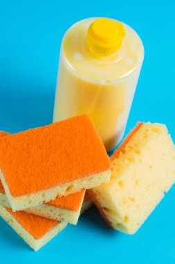 Şeffaf plastik bir şişede bulaşık deterjanı ve mavi arka planda gözenekli sarı temizlik süngeri. Ekolojik deterjan kavramı.