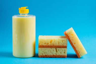 Şeffaf plastik bir şişede bulaşık deterjanı ve mavi arka planda gözenekli sarı temizlik süngeri. Ekolojik deterjan kavramı.