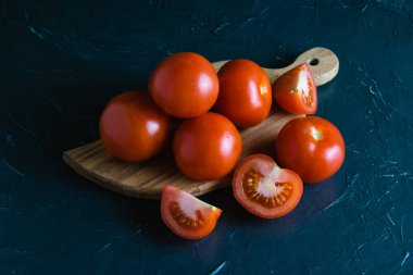 Bir sürü kırmızı domates, koyu beton arkaplanı ve fotokopi alanı ile parçalamak için ahşap bir tahtaya yakın duruyor..