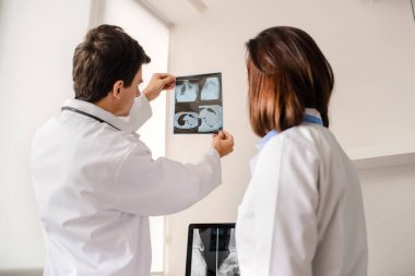 Tıp odasında iki genç doktor röntgen ve bilgisayar tomografisi filmlerini analiz ediyor ve defterde tıbbi bir açıklama yapıyor.