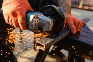 Çalışma sürecini kapatan koruyucu eldivenli işçi, elektrikli açı öğütücüyle metal bir çubuk kesiyor ve bir sürü kıvılcım çıkarıyor.