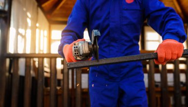 Çalışma sürecini kapatan koruyucu eldivenler ve mavi tulumlar giyen genç işçi metal bir çubuğu elektrik açı öğütücüsüyle kesiyor..