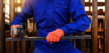 Çalışma sürecini kapatan koruyucu eldivenler ve mavi tulumlar giyen genç işçi metal bir çubuğu elektrik açı öğütücüsüyle kesiyor..