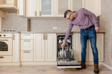 Genç adam bulaşıkları açık bulaşık makinesine koydu. Eski ahşap mutfakta temiz tabaklar vardı.