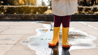 Yağmurdan sonra su birikintisinin ortasında duran sarı çizmeli çocuk bacaklarını kapat.