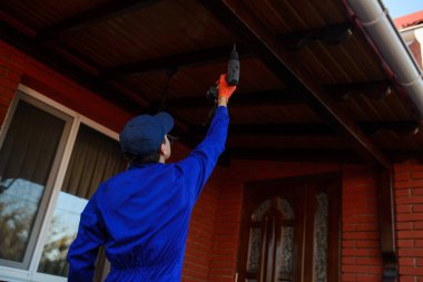 Mavi tulumlu ve koruyucu eldivenli genç müteahhit çatıdaki tahta çubuğu tamir ediyor.