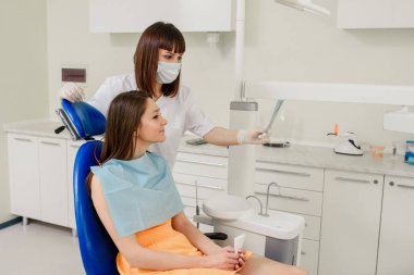 Genç kadın dişçi hastasına diş röntgeni panoramik radyografisinde diş hekimliğinde yapılması gereken tüm tedaviyi anlatıyor. Diş Xray dosyası tutuluyor