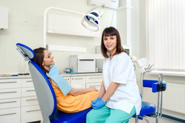 Diş kliniğinde gülümseyen genç dişçi kadın kavramı hastasının yanındaki kameraya bakıyor.