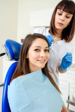 Dişçi kliniğinde gülümseyen genç bir kadın diş tedavisi görüyor ve kameraya bakıyor. Diş teçhizatlı dişçi arkada.