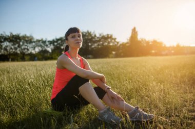 Genç ve güzel bir kadın spor yaptıktan ve gün batımında koştuktan sonra çimlerin üzerinde dinleniyor..
