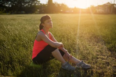 Genç ve güzel bir kadın spor yaptıktan ve gün batımında koştuktan sonra çimlerin üzerinde dinleniyor..