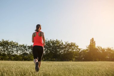 Genç bir kadının geriye bakış açısı spor yapıyor, gün batımında doğanın ortasında çimlerin üzerinde koşuyor. Her yerde spor yap.