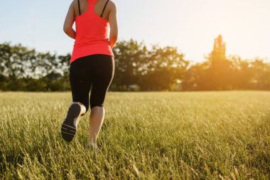 Genç bir kadının geriye bakış açısı spor yapıyor, gün batımında doğanın ortasında çimlerin üzerinde koşuyor. Her yerde spor yap.