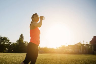 Genç ve güzel bir kadın gün batımında koşup spor yaptıktan sonra çimlerin üzerinde su içiyor.. 