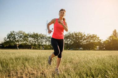 Genç ve güzel bir kadın gün batımında çimlerin üzerinde koşuyor. Her yerde spor yap.