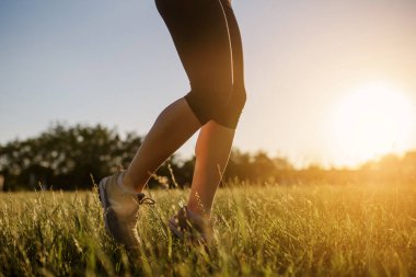 Spor yapan bir kadının bacaklarını kapayarak gün batımında doğanın ortasında çimlerin üzerinde koşuyor. Her yerde spor yap.