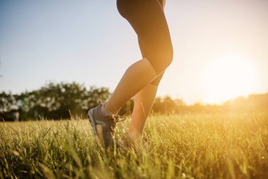 Spor yapan bir kadının bacaklarını kapayarak gün batımında doğanın ortasında çimlerin üzerinde koşuyor. Her yerde spor yap.