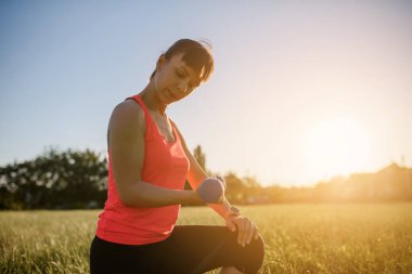 Genç ve güzel bir kadın spor yapıyor, gün batımında doğanın ortasında çimlerin üzerinde dambılla egzersiz yapıyor. Sonuçları analiz etmek için ukalalık taslıyor. Her yerde spor yap.