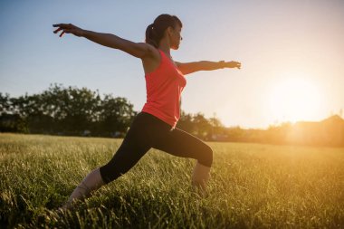 Genç ve güzel bir kadın spor yapıyor, gün batımında doğanın ortasında çimlerin üzerinde yoga egzersizleri yapıyor. Her yerde spor yap.