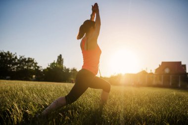 Genç ve güzel bir kadın spor yapıyor, gün batımında doğanın ortasında çimlerin üzerinde yoga egzersizleri yapıyor. Her yerde spor yap.