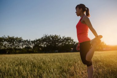 Genç ve güzel bir kadın spor yapıyor, gün batımında doğanın ortasında çimlerin üzerinde bacak egzersizleri yapıyor ve koşmaya hazırlanıyor. Her yerde spor yap.