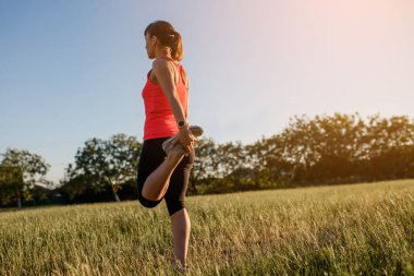Genç ve güzel bir kadın spor yapıyor, gün batımında doğanın ortasında çimlerin üzerinde bacak egzersizleri yapıyor ve koşmaya hazırlanıyor. Her yerde spor yap.