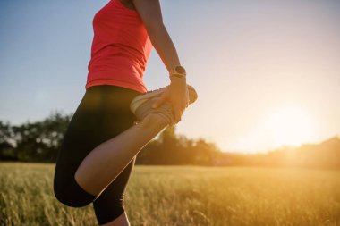 Genç ve güzel bir kadın spor yapıyor, gün batımında doğanın ortasında çimlerin üzerinde bacak egzersizleri yapıyor ve koşmaya hazırlanıyor. Her yerde spor yap.
