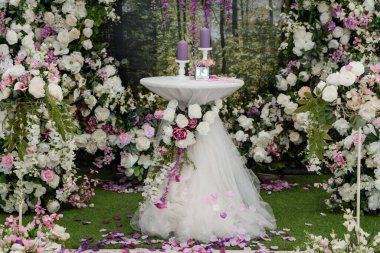 Düğün kemeri güllerle süslenmiş. Ortasında mum kompozisyonu olan bir masa var. Fotoğraf kabini bölgesi. Kimse yok.