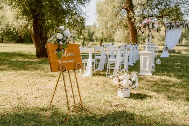 Hoş geldin standı, parkta düğün töreni. Güllerle süslenmiş beyaz sandalyeler. Kimse yok.