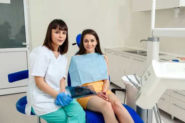 歯科医院の若い幸せな笑顔の歯科医の女性の概念は 彼女の美しい患者と一緒にカメラを見ており 歯科X線を保持しています 歯科パノラマX線 — ストック写真