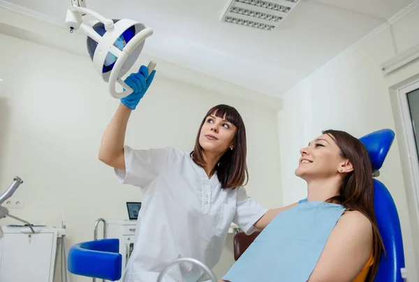 若い幸せな笑顔の女性歯科医は歯科の光を調整し 胃椅子に座っている患者の歯痛を治療する準備をしています — ストック写真