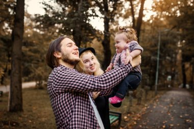Güzel, genç bir anne bebeğini elinde tutuyor ve babası yanında. Mutlu genç aile kavramı. Genç baba ve anne 6 aylık kızlarıyla parkta oynuyorlar.. 