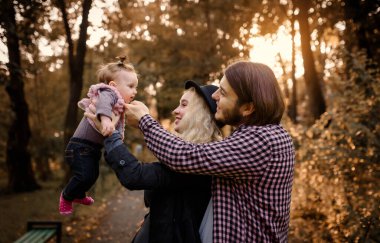 Güzel, genç bir anne bebeğini elinde tutuyor ve babası yanında. Mutlu genç aile kavramı. Genç baba ve anne 6 aylık kızlarıyla parkta oynuyorlar.. 