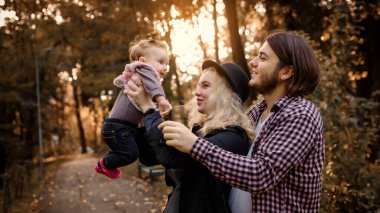 Güzel, genç bir anne bebeğini elinde tutuyor ve babası yanında. Mutlu genç aile kavramı. Genç baba ve anne 6 aylık kızlarıyla parkta oynuyorlar.. 