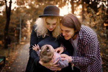 Güzel, genç bir anne bebeğini elinde tutuyor ve babası yanında. Mutlu genç aile kavramı. Genç baba ve anne 6 aylık kızlarıyla parkta oynuyorlar.. 
