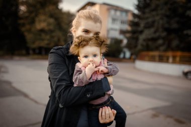 Genç anne, altı aylık bebeğini sonbahar parkında kollarında tutuyor. Küçük kız kameraya bakıyor..