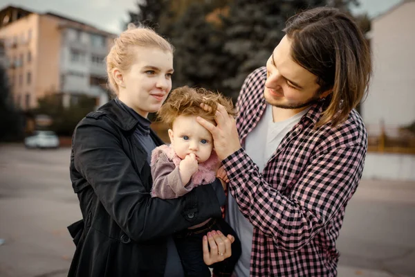 Genç anne ve baba, altı aylık kızlarını sonbahar parkında kollarında tutuyorlar. Küçük kız kameraya bakıyor..