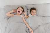 Ranní rodinný koncept. Vrchní pohled na bratra a sestru dítě v bílých tričkách probouzí a usmívá se do kamery v posteli