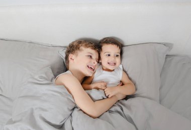 Erkek ve kız kardeş sabah kavramı. Küçük çocuk kız kardeşine yatakta sarılıyor ve uyandıktan sonra gülüyor. Beyaz tişört giyiyorlar.